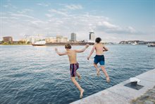 Twee kinderen springen in het water bij Amsterdam met boten en gebouwen op de achtergrond